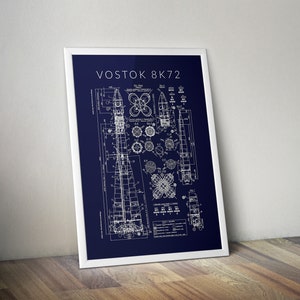 May include: A framed blueprint of the Vostok 8K72 rocket, displayed against a white background. The blueprint is in white on a dark blue background. The frame is white. The image is leaning against a wooden surface.