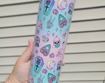 May include: A pastel blue and pink stainless steel tumbler with a variety of spooky and cute graphics, including a coffin, a crescent moon, an eye, a cupcake, a skull, and a peace sign. The text "Do Not Disturb" and "Goodbye" are also visible on the tumbler.