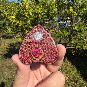 May include: A heart-shaped Ouija board with a glittery red surface. The board features gold lettering that spells out "YES", "NO", and "GOODBYE". A crescent moon and a circle with glitter are also visible. The board is held in a hand.