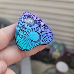 May include: A teal and purple planchette with the words "YES", "NO", and "GOODBYE" etched into the surface. The planchette has a crescent moon and a circular window filled with glitter. The planchette is a divination tool.