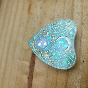May include: A blue and gold heart-shaped ouija board with the words "Hello", "Yes", "No", "Goodbye" and a crescent moon. The board is covered in glitter.