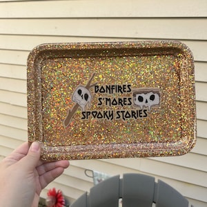 May include: A gold glitter tray with a sticker that says "Bonfires S'mores Spooky Stories". The sticker features two cartoon marshmallows with skulls for faces.