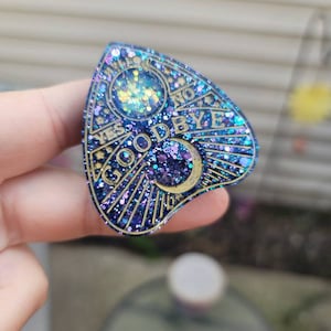May include: A glittery blue and purple planchette with gold accents. The planchette has the words "YES", "NO", and "GOODBYE" in gold lettering. A crescent moon and a circle are also visible.