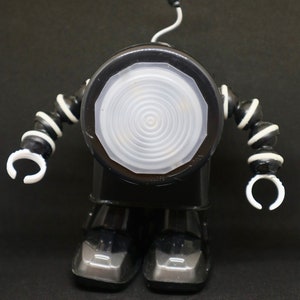 Puede incluir: Un juguete robot negro y blanco con una cabeza redonda, blanca y luminosa y brazos articulados. El robot tiene un cuerpo negro y detalles blancos en los brazos y las piernas.