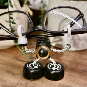 Puede incluir: Un pequeño robot caprichoso hecho de materiales reciclados. El robot lleva unas gafas con montura negra y tiene un ojo grande, redondo y con borde dorado. El robot está de pie sobre dos tapones de botella negros que dicen "Recíclame" en letras blancas.