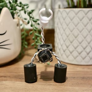 May include: A small, black, robot-like figure with a white ring on top and three black legs. The figure is made of metal and has a small, black camera lens in the center of its body.