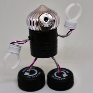 May include: A whimsical robot made from recycled materials. The robot has a clear plastic head with a silver eye, a black body, and white plastic arms. The robot is standing on two black bottle caps that say "Recycle Me".