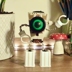May include: A white robot toy with a green eye, white arms, and white legs. The robot is standing on a wooden surface with two red and white mushroom figurines.