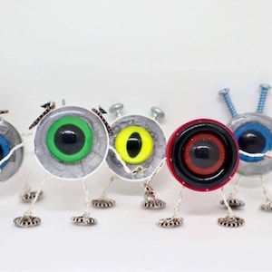 May include: Five small, whimsical robots made from recycled materials. Each robot has a different coloured eye, including blue, green, yellow, red, and black. The robots have silver bodies and are standing on a white surface.