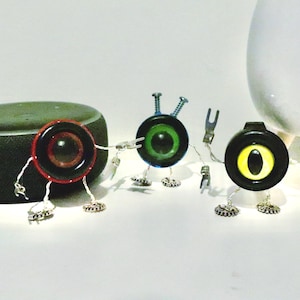 May include: A whimsical art piece featuring three robot-like figures with circular eye-like faces in red, green, and yellow. They have wire limbs and gear-shaped feet. A dark gray speaker and a white vase are in the background.