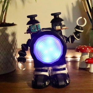 Puede incluir: Un robot negro hecho de materiales reciclados con una luz azul que brilla desde su centro. El robot tiene dos brazos, dos piernas y una cabeza con dos antenas.