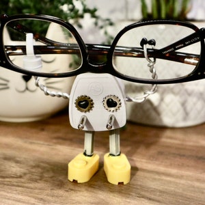May include: A white plastic figure with black heart-shaped eyes and gold gears, holding a pair of brown tortoiseshell eyeglasses. The figure has yellow plastic feet and is standing on a wooden surface.
