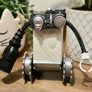 Könnte beinhalten: Ein weißer Roboter mit schwarzen Rädern und einem schwarzen Kabel. Der Roboter hat ein Bedienfeld mit Knöpfen und einem Drehknopf. Der Roboter steht auf einer Holzoberfläche.
