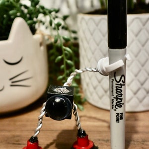 May include: A silver robot with red and black accents is standing on a wooden surface. The robot has a black cube for a head with a black eye. The robot is holding a black and white Sharpie fine point permanent marker.