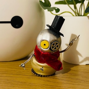 May include: A small, silver robot with a red scarf and a black top hat. The robot has one yellow eye and a gear on the side of its head. The robot is standing on a wooden surface.