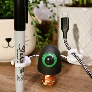 May include: A black and green robot made from a light bulb, wire, and a phone charger. The robot has a single green eye and is holding a black permanent marker with a white cap.
