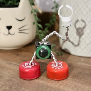 May include: A small robot made from recycled materials. The robot has a green eye, a black body, and red bottle caps for feet. The robot is attached to a white plastic ring.
