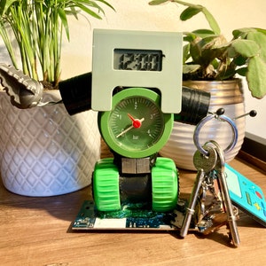 Puede incluir: Un reloj robótico verde y gris con una esfera redonda que indica "1 bar" y una pantalla digital que indica "12:00". El reloj está sobre una superficie de madera con un juego de llaves y un cartucho de juego azul y blanco.