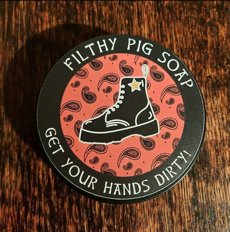 May include: A round black soap container with a red paisley pattern and a black boot with a gold star. The text "Filthy Pig Soap" and "Get Your Hands Dirty!" is printed in white around the edge of the container.