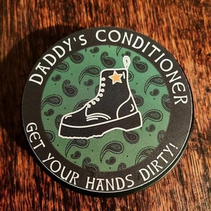 Könnte beinhalten: Ein runder schwarzer und grüner Aufkleber mit dem Text "Daddy's Conditioner Get Your Hands Dirty!" in Weiß. Ein schwarzer Stiefel mit einem weißen Stern an der Seite befindet sich in der Mitte des Aufklebers.