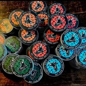 May include: A collection of round, colorful stickers with various designs featuring boots, text, and leopard print patterns. The stickers have black borders and white text that reads "Get Your Hands Dirty" and "Subby's Soft Soap".
