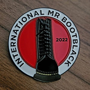 Könnte beinhalten: Eine schwarz-weiße Emaille-Anstecknadel mit rotem Hintergrund. Die Anstecknadel zeigt einen schwarzen Stiefel mit weißen Nähten und den Text "International Mr Bootblack 2022".