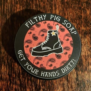 Könnte beinhalten: Ein runder schwarzer und roter Seifenbehälter mit dem Text "Filthy Pig Soap Get Your Hands Dirty!" und einem schwarzen Stiefel mit einem goldenen Stern.