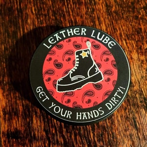 May include: A black and white sticker with a red paisley pattern and the text "Leather Lube Get Your Hands Dirty!"  A black boot with a white sole and a gold star is in the center of the sticker.