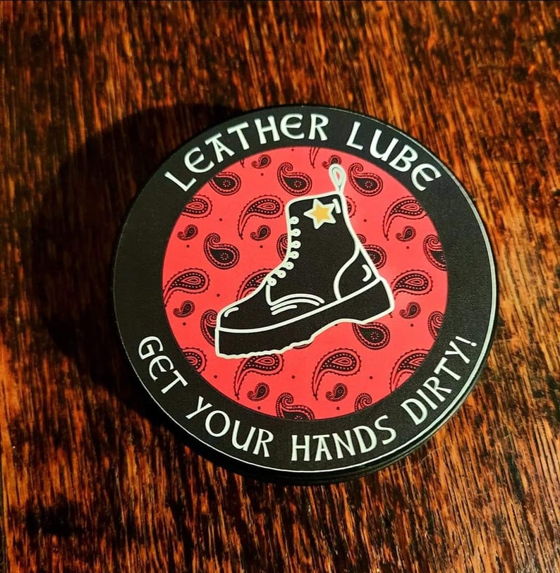 May include: A black and white sticker with a red paisley pattern and the text "Leather Lube Get Your Hands Dirty!"  A black boot with a white sole and a gold star is in the center of the sticker.