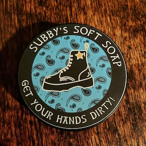 Könnte beinhalten: Ein runder Aufkleber in Schwarzweiß mit einem blauen Paisley-Muster. Der Aufkleber zeigt einen schwarzen Stiefel mit einem gelben Stern und dem Text "Subby's Soft Soap Get Your Hands Dirty!"