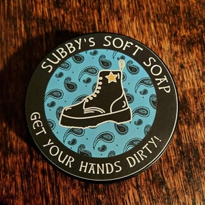 May include: A black and white circular sticker with a blue paisley pattern. The sticker features a black boot with a yellow star and the text "Subby's Soft Soap Get Your Hands Dirty!"