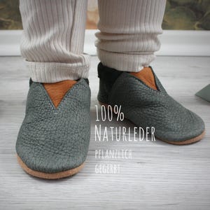 May include: A pair of gray leather baby shoes with brown leather accents. The shoes are made of 100% natural leather and are plant-tanned. The text "100% NATURLEDER PFLANZLICH GEGERBT" is printed on the shoes.