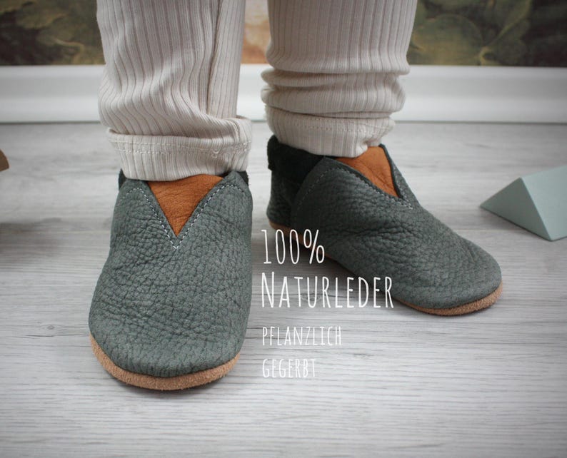 May include: A pair of gray leather baby shoes with brown leather accents. The shoes are made of 100% natural leather and are plant-tanned. The text "100% NATURLEDER PFLANZLICH GEGERBT" is printed on the shoes.