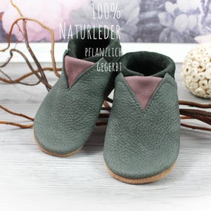 May include: A pair of dark green leather baby shoes with a pink leather trim. The shoes are made of 100% natural leather and are vegetable tanned. The text "100% NATURLEDER PFLANZLICH GEGERBT" is visible on the image.