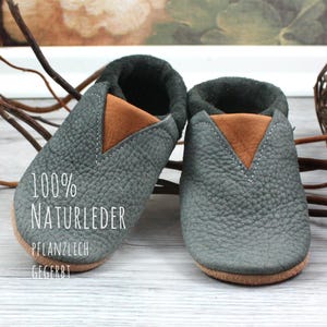 May include: A pair of gray leather baby shoes with brown leather accents. The shoes are made of 100% natural leather and are vegetable tanned. The text "100% NATURLEDER PFLANZLICH GEGERBT" is printed on the sole of one shoe.