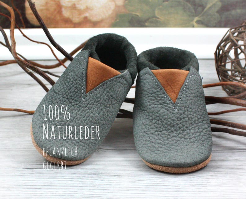 May include: A pair of gray leather baby shoes with brown leather accents. The shoes are made of 100% natural leather and are vegetable tanned. The text "100% NATURLEDER PFLANZLICH GEGERBT" is printed on the sole of one shoe.