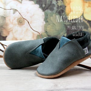 May include: A pair of gray leather baby shoes with a blue suede trim. The shoes are made of 100% natural leather and are plant-tanned. The text "100% NATURLEDER PFLANZLICH GEGERBT" is visible on the background.