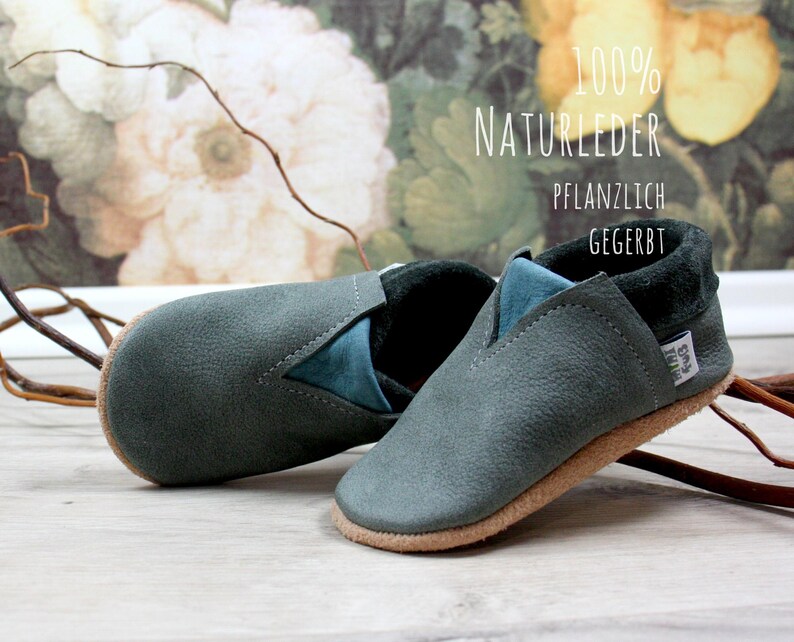 May include: A pair of gray leather baby shoes with a blue suede trim. The shoes are made of 100% natural leather and are plant-tanned. The text "100% NATURLEDER PFLANZLICH GEGERBT" is visible on the background.