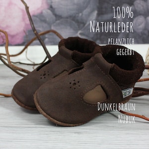 May include: A pair of dark brown leather baby shoes with a velcro strap closure. The shoes are made of 100% natural leather and are vegetable tanned. The text "100% NATURLEDER", "PFANZLICH GEGERBT", "DUNKELBRAUN NUBUK" is printed on the shoes.