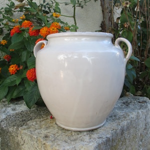 Op de afbeelding: Een glanzende, gebroken witte keramische pot met een rond lichaam, een brede ronde opening en twee kleine, gebogen handvatten aan weerszijden bij de rand. Het oppervlak van de pot is glad en reflecteert het licht op een egale wijze.