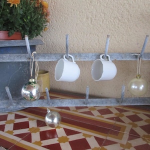 Op de afbeelding: Een rustieke mokkenrek van gegalvaniseerd metaal met twee witte mokken eraan. De rek heeft ook twee kerstversieringen. Een plant in een pot en een gele container staan op de achtergrond. De rek staat op een betegeld oppervlak.