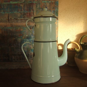 May include: A light blue enamel coffee pot with a black trim and a spout. The pot is sitting on a wooden table with two other ceramic pots.
