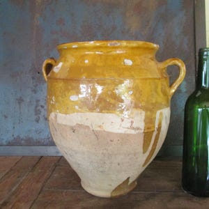 Puede incluir: Una gran jarra de cerámica antigua de color amarillo y crema con dos asas. La jarra tiene una boca ancha y un cuerpo bulboso. Dos botellas de vino verdes están en el fondo. La jarra probablemente se utiliza para almacenamiento o decoración.