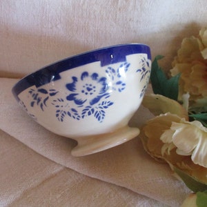 May include: A small, white ceramic bowl with a blue floral design and a blue rim. The bowl has a pedestal base and is set against a neutral background with floral accents.