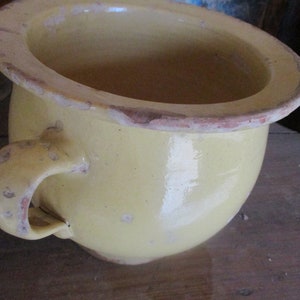 Pode incluir: Um pote de cerâmica amarelo com uma borda larga e uma única alça. O pote tem um aspecto desgastado e rústico e é envidraçado em uma cor amarelo pálido.