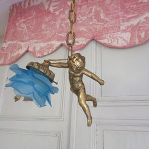Puede incluir: Una figurita de querubín dorada sosteniendo una pantalla de lámpara de vidrio azul. El querubín está suspendido de una cadena y está en posición de vuelo.