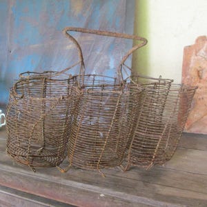 May include: A vintage, rustic metal wire basket with three compartments and a handle. The basket is made of rusty brown wire and sits on a wooden surface. The background includes a blue and yellow wall and a terracotta sculpture.