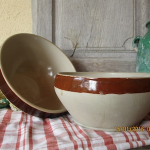 May include: Two ceramic bowls with brown rims. The larger bowl is beige and the smaller bowl is white. Both bowls are sitting on a red and white checkered tablecloth.