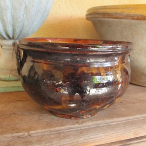Puede incluir: Una olla de cerámica marrón glaseada con dos asas. La olla tiene un esmalte marrón oscuro con un ligero brillo. La olla está sentada sobre una superficie de madera.