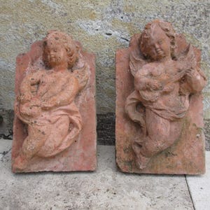 Peut inclure: Deux sculptures de chérubins en terre cuite, chacune représentant un ange ailé avec un instrument de musique. Les sculptures sont montées sur des carreaux rectangulaires.
